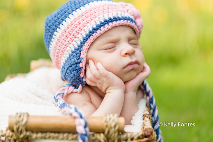 Fotografia Newborn RJ Recém Nascido Bebê com touca azul com tranças flor rosa segurando cabeça sobre as mãos dentro cestinha por Kelly Fontes